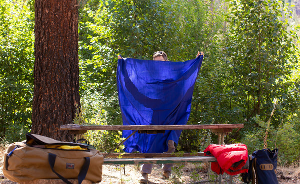 A Large Shepherd's Wool Utility Cloth in Neela Blue being shaken into the air to spread as a table cloth on a picnic table at a campsite.